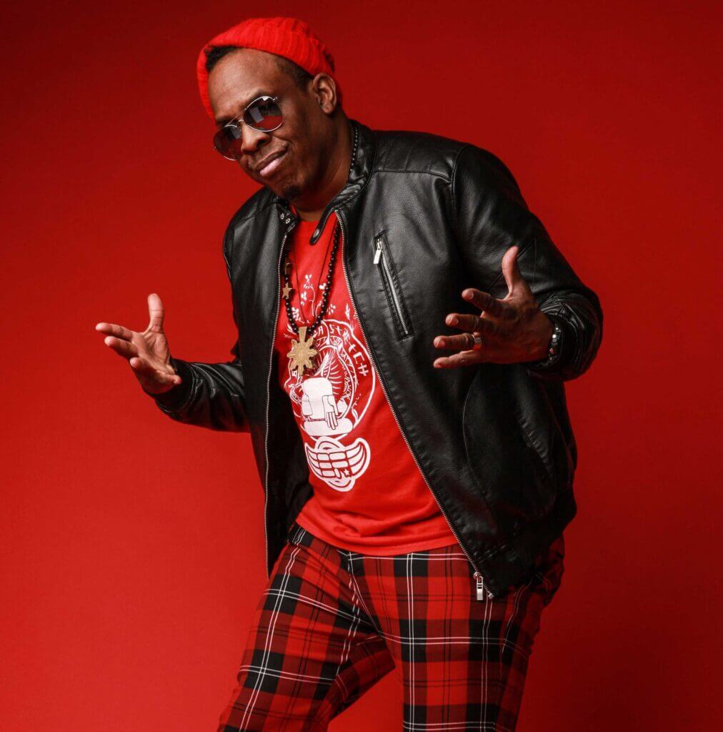 Photo of Buddha Stretch with red background, red plaid pants, red tee shirt, red hat, and black leather jacket.